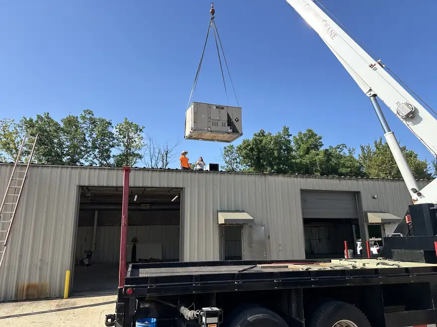A crane lifting a new rooftop HVAC unit onto a commercial metal building